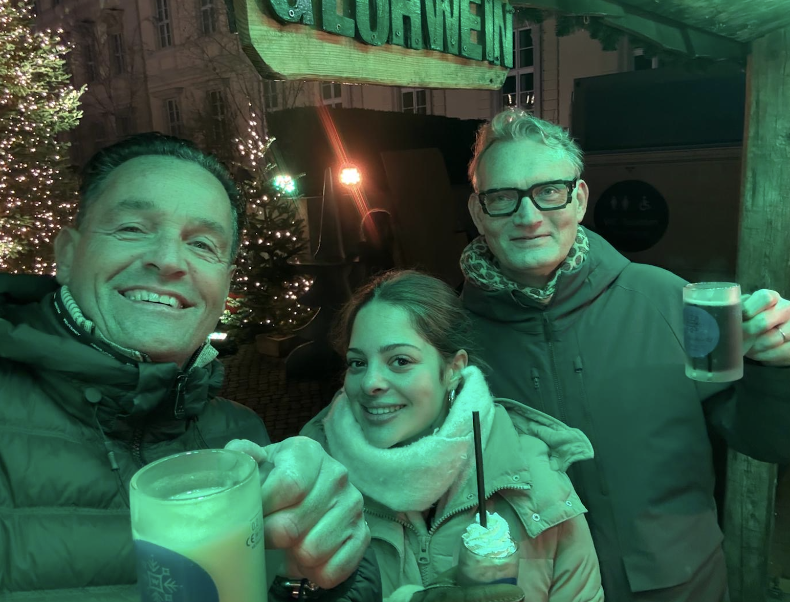 Drie personen staan buiten bij een verlichte Gl&uuml;hwein-stand en houden warme drankjes vast.